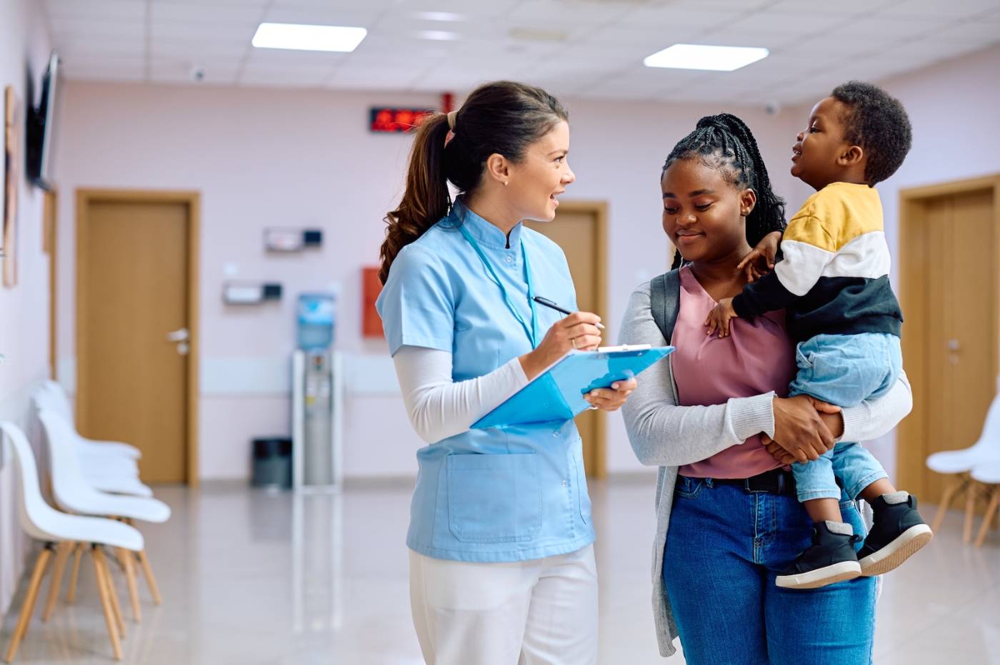 Female medical assistant with clipboard talks with a young mother holding toddler